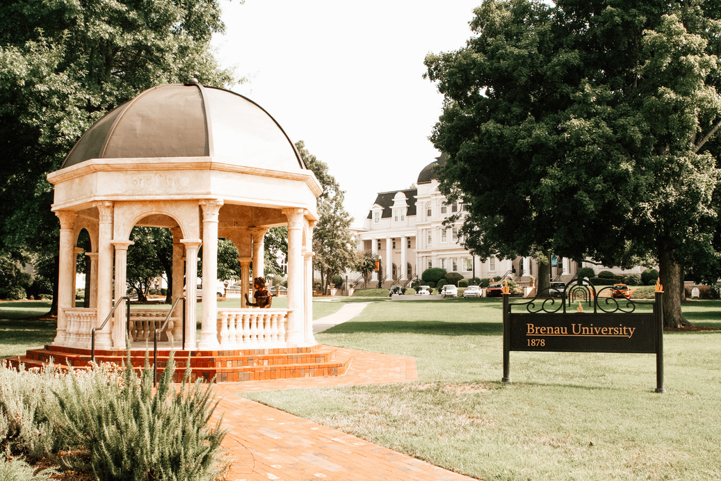 [Alt-text]: a gazebo sits in the middle of a courtyard in front of large white university building. Sign runs outside of frame reading ‘Brenau University 1876’