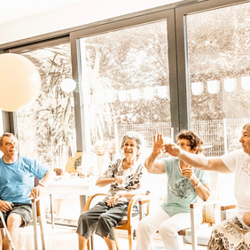 [Alt-text]: a group of older adults sitting in a room tossing a balloon in the air
