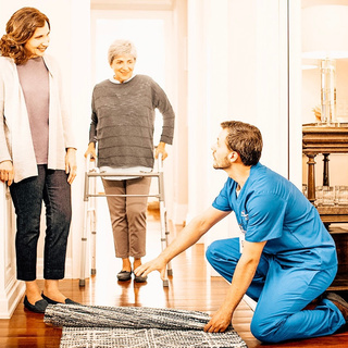[Alt-text]: a person in blue scrubs is helping an elderly person with a walker by removing rug to reduce fall risk 